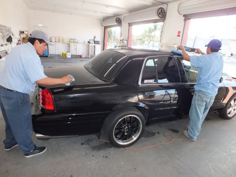 2003 Ford Crown Victoria - Black sedan rear three-quarter with one worker buffing trunk and another applying tint film.