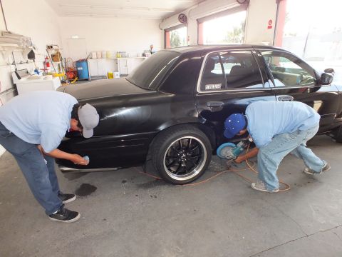 2003 Ford Crown Victoria - Black sedan side profile with techs polishing the lower body and quarter panel in a shop bay.
