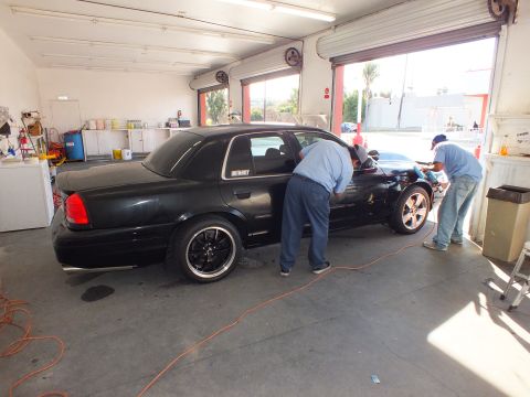 2003 Ford Crown Victoria - Rear three-quarter of black sedan on black wheels with two techs working at the front end.