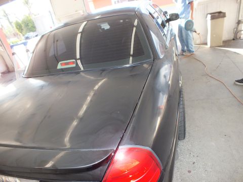 2003 Ford Crown Victoria - Technician with a spray applicator behind a dark sedan whose decklid shows buffing swirls.