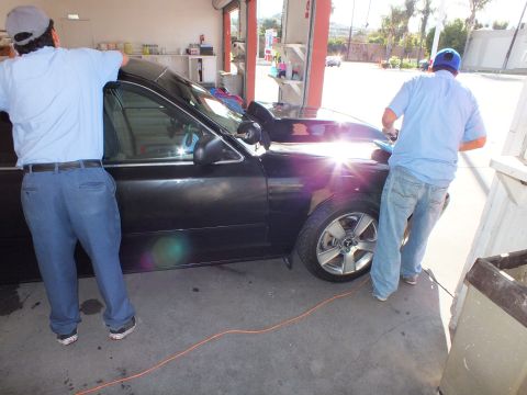 2003 Ford Crown Victoria - Two workers wipe down the front of a black sedan in a sunlit detail shop bay.