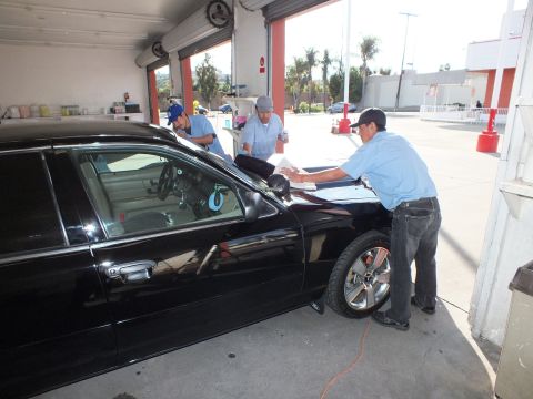2003 Ford Crown Victoria - Three detailers wipe the windshield and front of a black sedan in a garage bay.