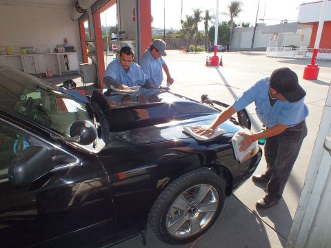 2003 Ford Crown Victoria - Detailers towel the hood and glass of a black sedan under bright daylight in a bay.