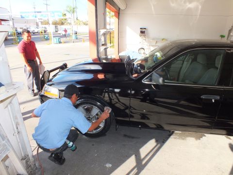 2003 Ford Crown Victoria - technician kneels to clean the front wheel of a black sedan parked in an open shop bay.