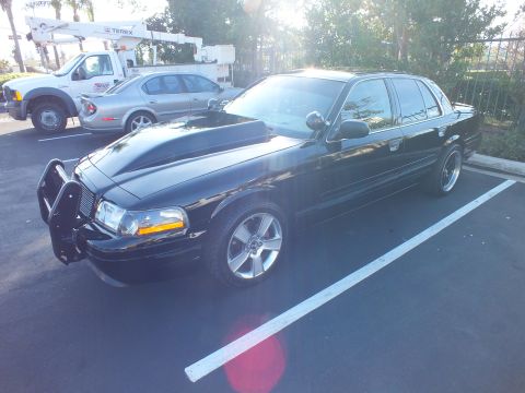 2003 Ford Crown Victoria - Modified black sedan parked by utility trucks showing raised hood and grille guard