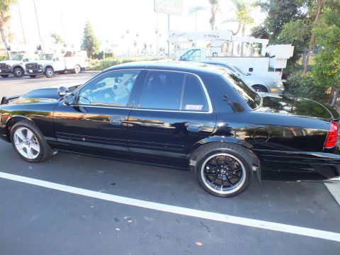 2003 Ford Crown Victoria - Rear quarter profile of a black full-size sedan parked beside work trucks