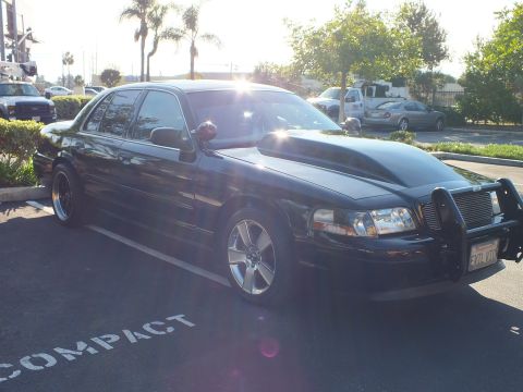2003 Ford Crown Victoria - Lowered black Mercury sedan, sun flaring over the hood, parked in a compact stall with a billet grille guard up front.