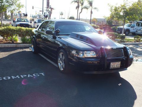 2003 Ford Crown Victoria - Front-end view of a lowered black sedan wearing a billet grille guard, palm trees and work trucks behind it.