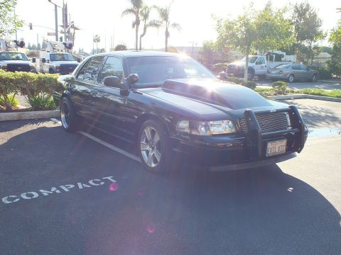 2003 Ford Crown Victoria - Sun-washed front shot of a blacked-out Mercury sedan with chrome grille guard, parked over a COMPACT stall marking.