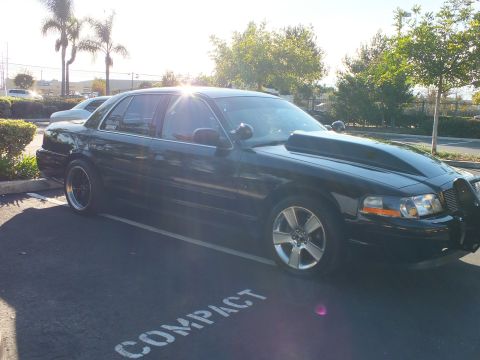 2003 Ford Crown Victoria - Low black sedan catches sun across the windshield, billet grille and amber side marker visible at the nose.