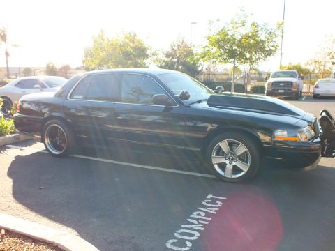 2003 Ford Crown Victoria - Lowered black sedan parked diagonally, chrome grille guard and amber marker at the front, pickups in the lot.