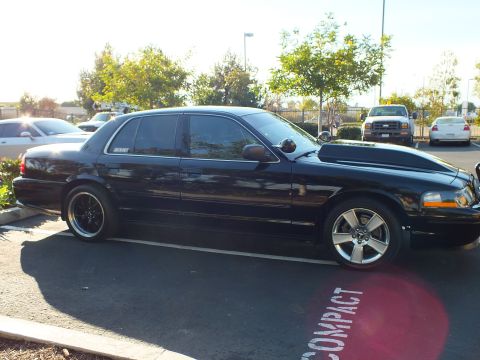 2003 Ford Crown Victoria - Side view of a dropped black full-size sedan; trees and parked pickups fill the background.