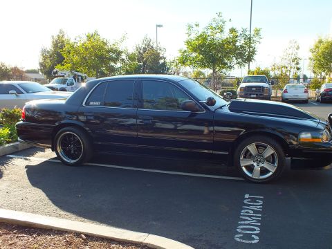 2003 Ford Crown Victoria - Glossy black full-size sedan sits low across the parking line, amber marker and grille guard up front.