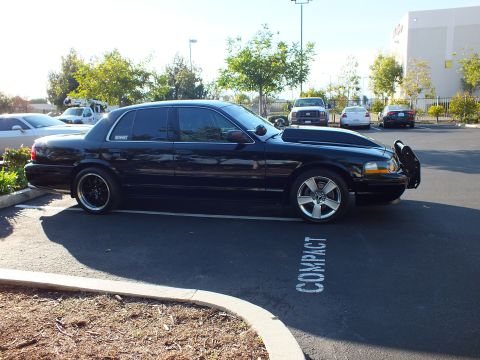2003 Ford Crown Victoria - Sun-lit passenger-side view of a dropped black sedan with chrome grille guard over a COMPACT stall.