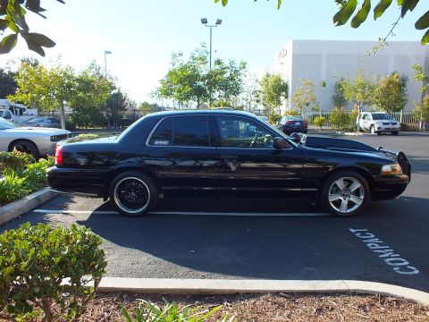 2003 Ford Crown Victoria - Clean passenger-side profile of a black full-size sedan parked under bright sun by a white warehouse.