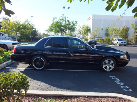 2003 Ford Crown Victoria - Black sedan in profile, amber side marker and grille guard at the front, warehouse and trees behind.