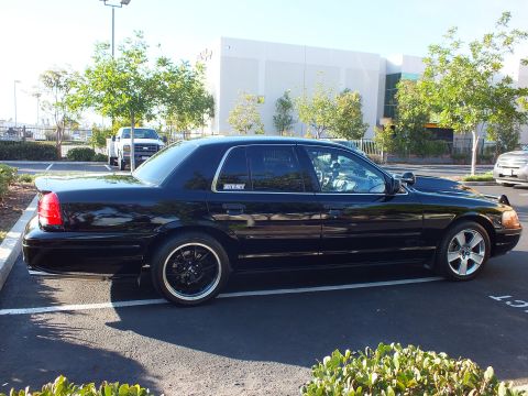 2003 Ford Crown Victoria - Rear-quarter view of a dropped black full-size sedan parked by a white warehouse, trees lining the lot.