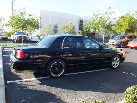 2003 Ford Crown Victoria - Dropped black sedan viewed from the rear quarter, white warehouse and parked cars filling the background.