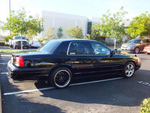 2003 Ford Crown Victoria - Glossy black sedan from the rear quarter, sun reflecting off the trunk lid in a sunny parking lot.