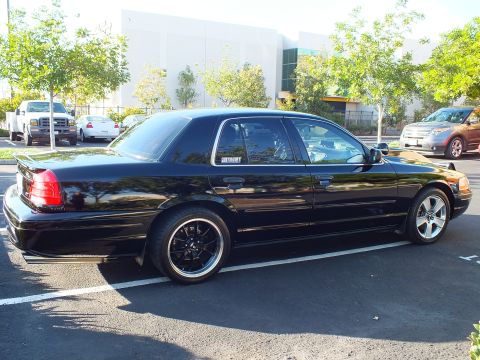 2003 Ford Crown Victoria - Side-rear view of a low glossy black sedan, MERCURY decal visible, parked among trucks and trees.