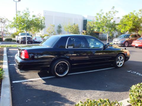 2003 Ford Crown Victoria - Rear three-quarter of a blacked-out full-size sedan sitting low, warehouse and parked cars behind it.