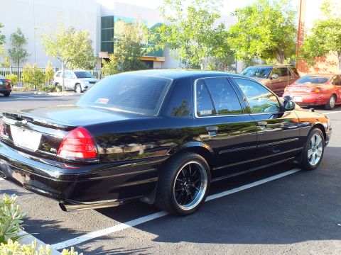 2003 Ford Crown Victoria - Lowered black Crown Victoria with dark spoke wheels parked beside shrubs and trees.