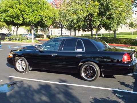 2003 Ford Crown Victoria - Black Crown Victoria sedan in profile on wet asphalt, dark spoke wheels.