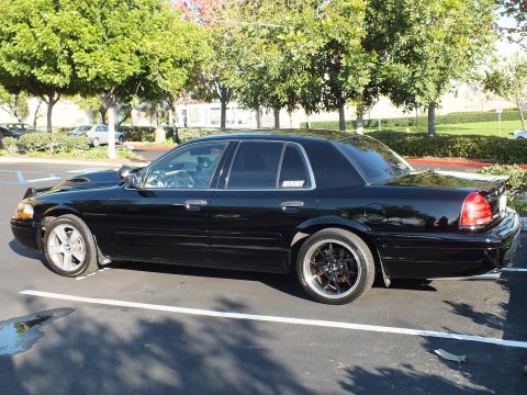 2003 Ford Crown Victoria - Lowered black Crown Victoria parked left-side-on, trees lining the lot behind.