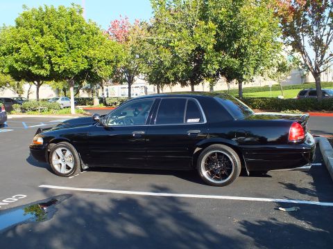 2003 Ford Crown Victoria - Black Crown Victoria sedan viewed from the side, casting a shadow across the asphalt.
