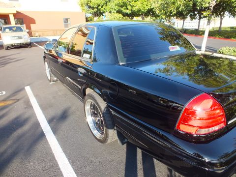 2003 Ford Crown Victoria - Blacked-out full-size sedan parked in a striped lot, glossy paint reflecting trees and sky.