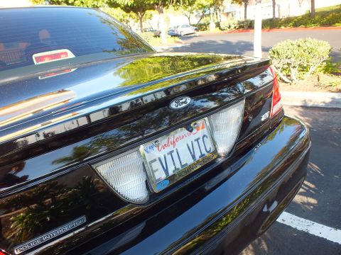 2003 Ford Crown Victoria - Sunlit black Ford decklid in a lot, showing EVIL VIC plate, Ford oval and Police Interceptor lettering.