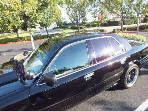 2003 Ford Crown Victoria - Side view of a black Ford sedan, dark tinted glass and a black aftermarket wheel catching light.