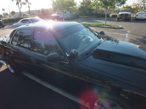 2003 Ford Crown Victoria - Backlit side-front shot of a black Ford sedan, sun flare over the windshield and door mirror.