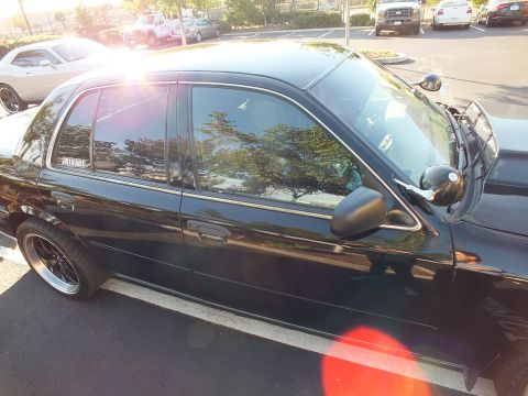 2003 Ford Crown Victoria - Sunlit driver-side view of a black Ford sedan with tinted glass and a white quarter-window decal.