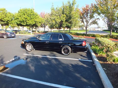 2003 Ford Crown Victoria - Wide side shot of a black Ford sedan with lowered stance and black wheels in a tree-lined lot.