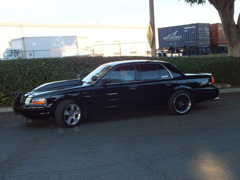 2003 Ford Crown Victoria - Black Ford sedan side view with push bar and black wheels parked near freight trucks and containers.