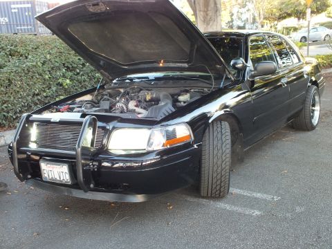 2003 Ford Crown Victoria - Engine bay exposed on a black Crown Vic, V8 visible behind a billet grille and front push bar, EVIL VIC plate.