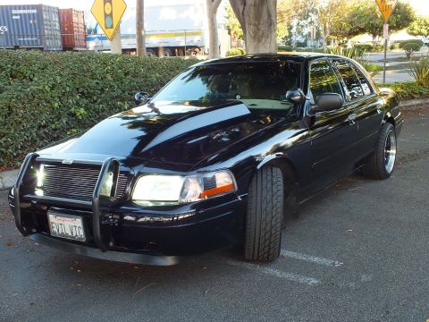 2003 Ford Crown Victoria - Front three-quarter of a black Crown Vic with lights lit, billet grille and chrome push bar, parked curbside.