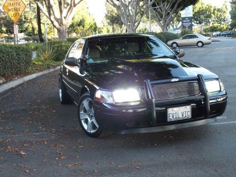 2003 Ford Crown Victoria - Three-quarter front of a dark Crown Vic with lit headlights and chrome push bar, EVIL VIC plate.