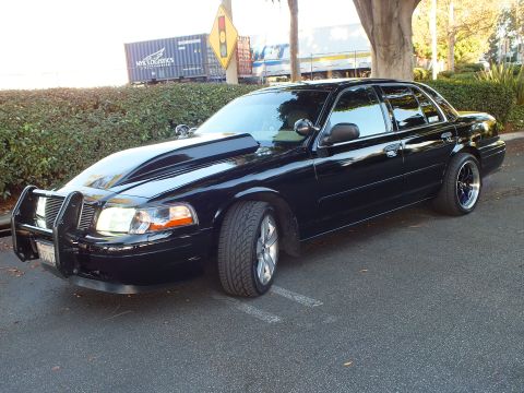 2003 Ford Crown Victoria - Profile-leaning shot of a black Crown Vic with machined wheels and a chrome grille guard, hedge backdrop.