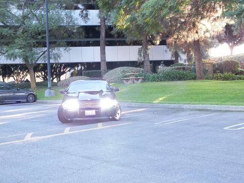 2003 Ford Crown Victoria - Distant view of a dark sedan with headlights on across an empty parking lot, office building behind.