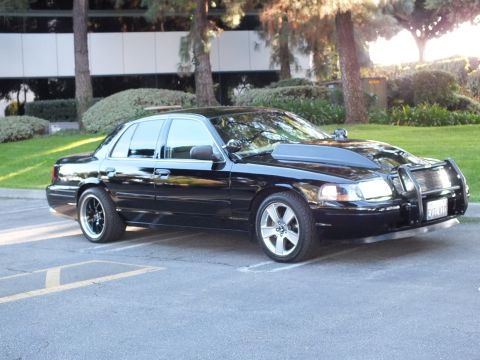 2003 Ford Crown Victoria - Black Crown Vic with chrome push bar and machined wheels, office building and greenery in back.