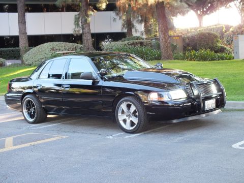 2003 Ford Crown Victoria - Black Crown Vic at an angle on machined wheels, billet grille behind a chrome guard, green lawn behind.