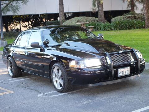 2003 Ford Crown Victoria - Black Crown Vic with headlights lit, billet grille behind a chrome push bar, EVIL VIC plate, lawn behind.