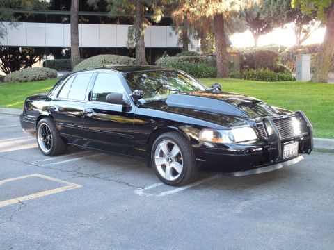 2003 Ford Crown Victoria - Black Crown Vic angled on machined wheels with a chrome guard, sun-lit office lawn in the background.