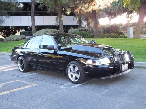 2003 Ford Crown Victoria - Modified black Ford Crown Victoria, front-three-quarter view, brush guard and aftermarket wheels, sunlit parking lot.