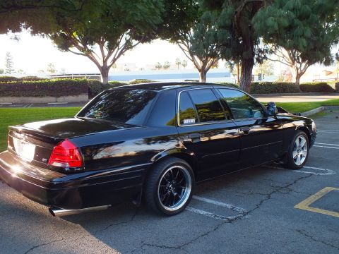 2003 Ford Crown Victoria - Rear-three-quarter view of lowered black Crown Victoria, dark wheels and chrome dual tips, trees behind.