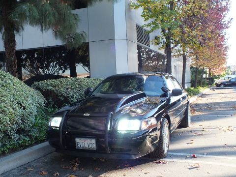 2003 Ford Crown Victoria - Dark Crown Victoria front quarter with lit headlights and brush guard, EVIL VIC plate, building behind.