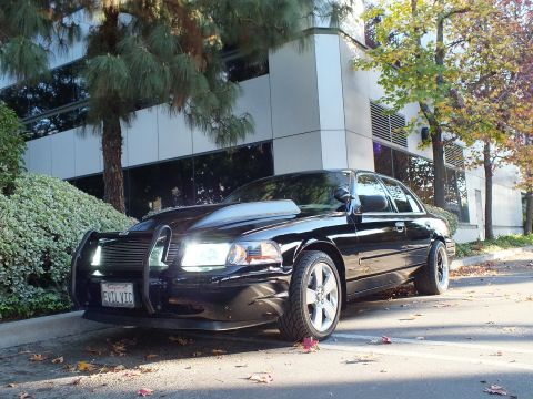 2003 Ford Crown Victoria - Front-three-quarter of black Crown Victoria with lit headlights and brush guard among fallen leaves.