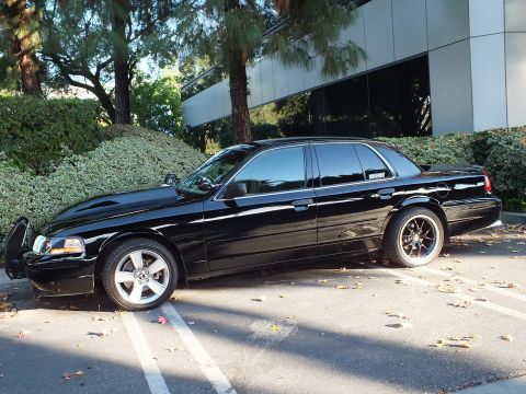 2003 Ford Crown Victoria - Full driver-side profile of a lowered black sedan on multi-spoke wheels, parked beside a glass office facade.